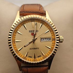 Op de afbeelding: Een goudkleurig Seiko automatisch polshorloge met een bruine leren band. De wijzerplaat heeft gouden wijzers en markeringen, met een dag-datumweergave op 3 uur. De achtergrond is neutraal.