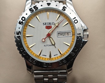May include: A silver Seiko 5 automatic wristwatch with a white dial and gold-colored accents. The watch features a day and date display, a silver band, and a rotating bezel with black numerals.