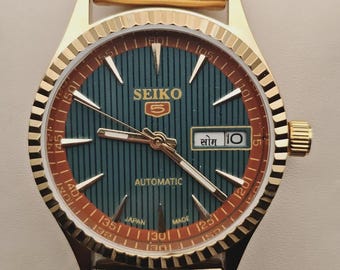 May include: A gold-tone Seiko 5 automatic wristwatch featuring a dark green vertical-striped dial with gold-tone indices and hands. The watch includes a day-date window at the three o'clock position, a fluted bezel, and a matching gold-tone metal link bracelet.