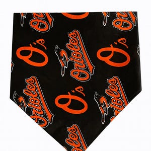 Op de afbeelding: Zwarte bandana met een all-over patroon van het Baltimore Orioles teamlogo en het woord "Orioles" in oranje en wit. De bandana heeft een driehoekige vorm.