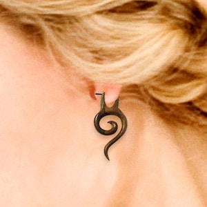 May include: A dark brown wooden earring with a spiral design. The earring is worn in the ear of a person with blonde hair.