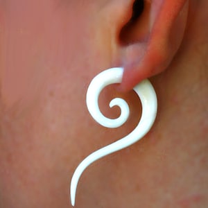 May include: White spiral ear gauge made of bone. The gauge is worn in a person's earlobe.