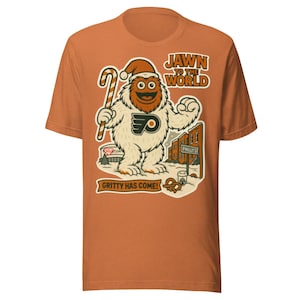 Op de afbeelding: Oranje t-shirt met een cartoon illustratie van Gritty, de mascotte van de Philadelphia Flyers, met een kerstmuts en een zuurstok. De tekst bevat "JAWN TO THE WORLD" en "GRITTY HAS COME!"