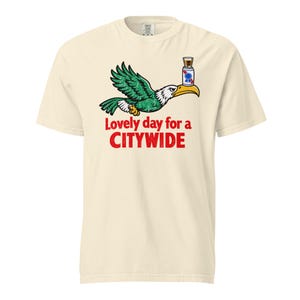 Può includere: Una t-shirt a maniche corte color crema con la grafica di un'aquila verde e bianca in volo che bilancia una lattina di birra sulla testa. Sotto l'aquila, il testo rosso recita "Lovely day for a CITYWIDE" in un carattere grassetto usurato.