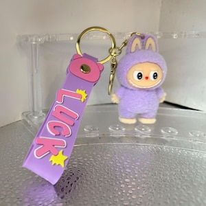 Peut inclure: Un porte-cl&eacute;s violet avec un anneau dor&eacute; et un personnage de dessin anim&eacute; en costume de lapin. La lani&egrave;re du porte-cl&eacute;s porte le mot "LUCK" en rose avec des &eacute;toiles jaunes. Le personnage a un visage souriant.