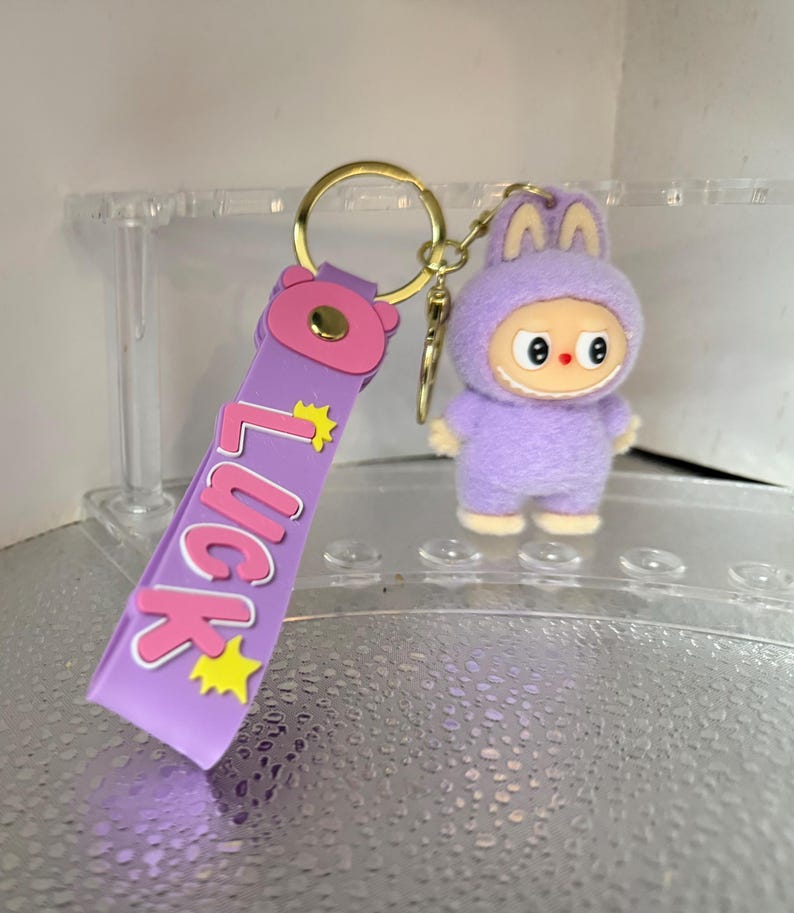 Peut inclure: Un porte-cl&eacute;s violet avec un anneau dor&eacute; et un personnage de dessin anim&eacute; en costume de lapin. La lani&egrave;re du porte-cl&eacute;s porte le mot "LUCK" en rose avec des &eacute;toiles jaunes. Le personnage a un visage souriant.