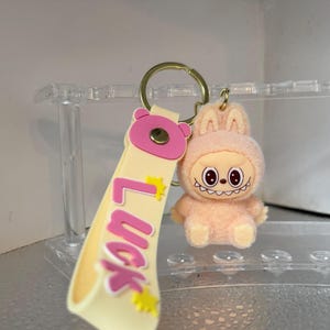 Peut inclure: Un porte-cl&eacute;s avec un personnage de lapin en peluche p&ecirc;che, souriant, et une lani&egrave;re assortie avec le mot "Luck" en lettres roses. Le porte-cl&eacute;s a un anneau de couleur or.