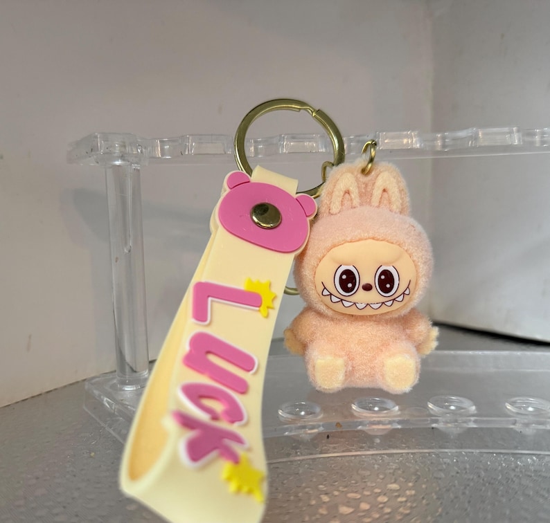 Peut inclure: Un porte-cl&eacute;s avec un personnage de lapin en peluche p&ecirc;che, souriant, et une lani&egrave;re assortie avec le mot "Luck" en lettres roses. Le porte-cl&eacute;s a un anneau de couleur or.