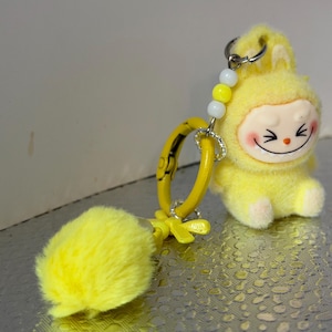 Peut inclure: Un porte-cl&eacute;s en peluche jaune repr&eacute;sentant un personnage de dessin anim&eacute; souriant avec des joues roses et une expression joyeuse. Le porte-cl&eacute;s comprend un anneau jaune, un pompon duveteux et un accent de perles. Le personnage porte une capuche.