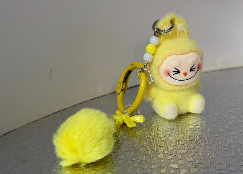 Peut inclure: Un porte-cl&eacute;s en peluche jaune repr&eacute;sentant un personnage de dessin anim&eacute; souriant avec des joues roses et une expression joyeuse. Le porte-cl&eacute;s comprend un anneau jaune, un pompon duveteux et un accent de perles. Le personnage porte une capuche.