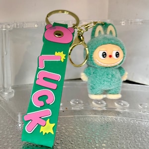 Peut inclure: Un porte-cl&eacute;s vert et rose avec le mot "Luck" et un personnage de dessin anim&eacute; dans un costume vert flou. Le porte-cl&eacute;s a un anneau et un fermoir dor&eacute;s.