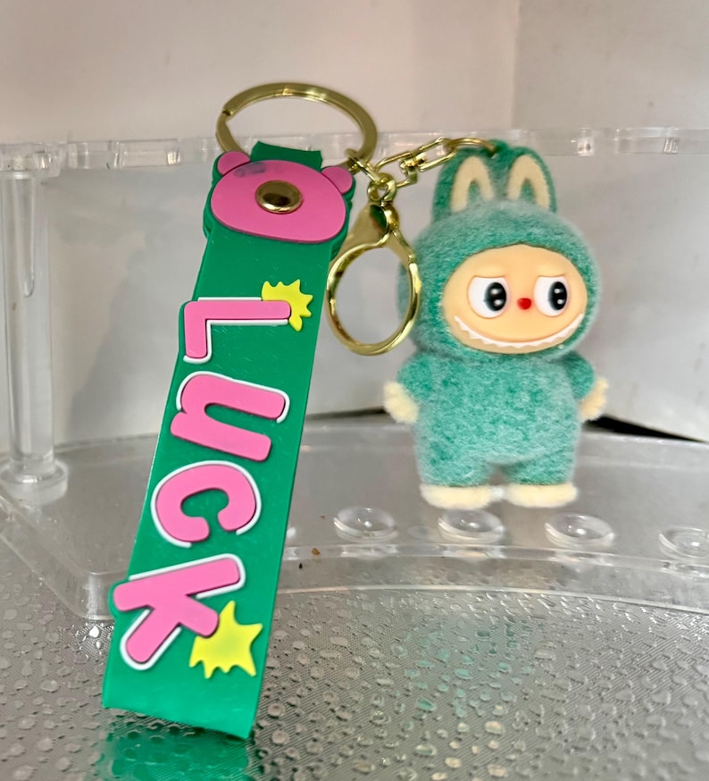 Peut inclure: Un porte-cl&eacute;s vert et rose avec le mot "Luck" et un personnage de dessin anim&eacute; dans un costume vert flou. Le porte-cl&eacute;s a un anneau et un fermoir dor&eacute;s.