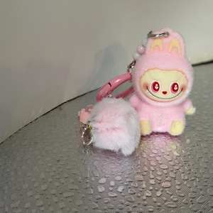Peut inclure: Un porte-cl&eacute;s en peluche rose repr&eacute;sentant un personnage de dessin anim&eacute; en costume de lapin avec un visage souriant et des yeux rouges. Le porte-cl&eacute;s comprend un pompon rose duveteux et un fermoir rose.