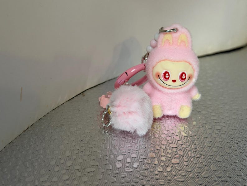 Peut inclure: Un porte-cl&eacute;s en peluche rose repr&eacute;sentant un personnage de dessin anim&eacute; en costume de lapin avec un visage souriant et des yeux rouges. Le porte-cl&eacute;s comprend un pompon rose duveteux et un fermoir rose.