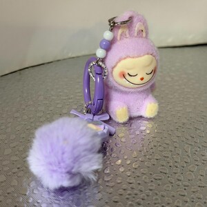 Peut inclure: Un porte-cl&eacute;s en forme de lapin en peluche lavande avec un visage souriant et les yeux ferm&eacute;s. Le porte-cl&eacute;s a un fermoir violet, un pompon duveteux et un accent perl&eacute;. Le lapin porte une tenue &agrave; capuche avec des pieds jaunes.