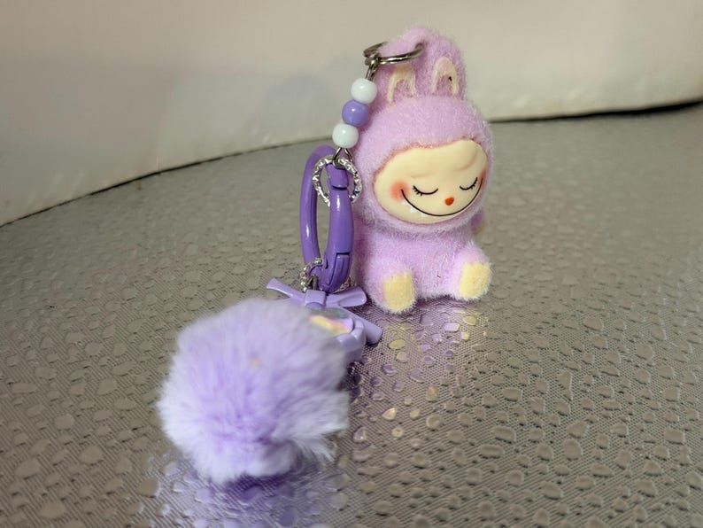 Peut inclure: Un porte-cl&eacute;s en forme de lapin en peluche lavande avec un visage souriant et les yeux ferm&eacute;s. Le porte-cl&eacute;s a un fermoir violet, un pompon duveteux et un accent perl&eacute;. Le lapin porte une tenue &agrave; capuche avec des pieds jaunes.