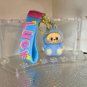 Peut inclure: Un porte-cl&eacute;s bleu avec le mot "Luck" en lettres roses et des &eacute;toiles jaunes. Une petite figurine en peluche bleue avec un visage souriant et un porte-cl&eacute;s dor&eacute; est attach&eacute;e. Le porte-cl&eacute;s est expos&eacute; sur un pr&eacute;sentoir en plastique transparent.