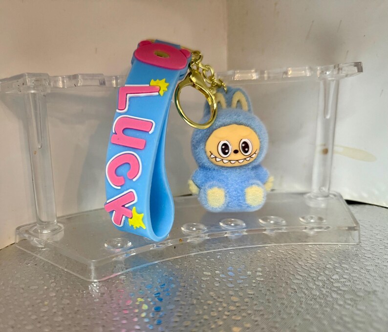 Peut inclure: Un porte-cl&eacute;s bleu avec le mot "Luck" en lettres roses et des &eacute;toiles jaunes. Une petite figurine en peluche bleue avec un visage souriant et un porte-cl&eacute;s dor&eacute; est attach&eacute;e. Le porte-cl&eacute;s est expos&eacute; sur un pr&eacute;sentoir en plastique transparent.