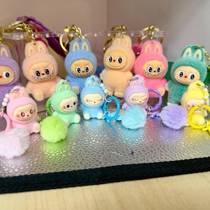 Peut inclure: Collection de porte-cl&eacute;s en peluche en forme de lapin. Chaque porte-cl&eacute;s pr&eacute;sente un personnage de lapin dans diff&eacute;rentes couleurs pastel, notamment le violet, le rose, le bleu et le jaune. Les lapins ont des traits de visage distinctifs et sont attach&eacute;s &agrave; des porte-cl&eacute;s dor&eacute;s avec des pompons.