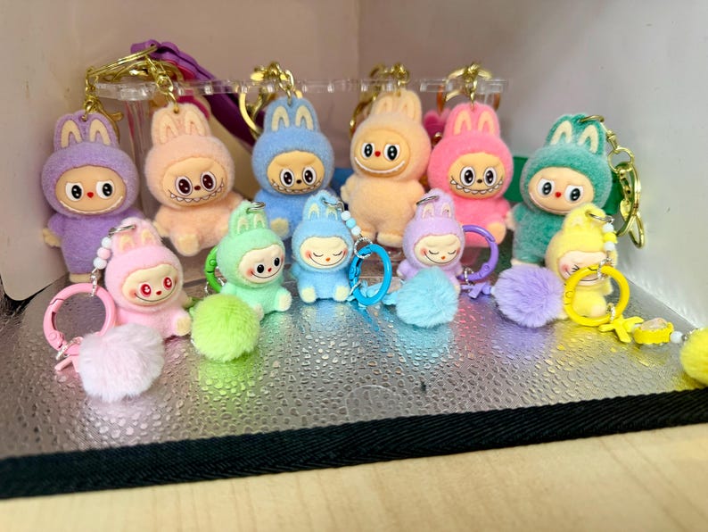 Peut inclure: Collection de porte-cl&eacute;s en peluche en forme de lapin. Chaque porte-cl&eacute;s pr&eacute;sente un personnage de lapin dans diff&eacute;rentes couleurs pastel, notamment le violet, le rose, le bleu et le jaune. Les lapins ont des traits de visage distinctifs et sont attach&eacute;s &agrave; des porte-cl&eacute;s dor&eacute;s avec des pompons.