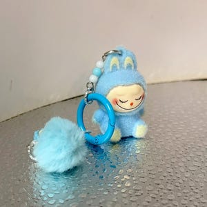 Peut inclure: Un porte-cl&eacute;s en peluche bleu clair repr&eacute;sentant un personnage de dessin anim&eacute; en costume de lapin. Le personnage a un visage souriant avec les yeux ferm&eacute;s et des accents jaunes. Un pompon bleu duveteux et un anneau bleu sont attach&eacute;s au porte-cl&eacute;s.