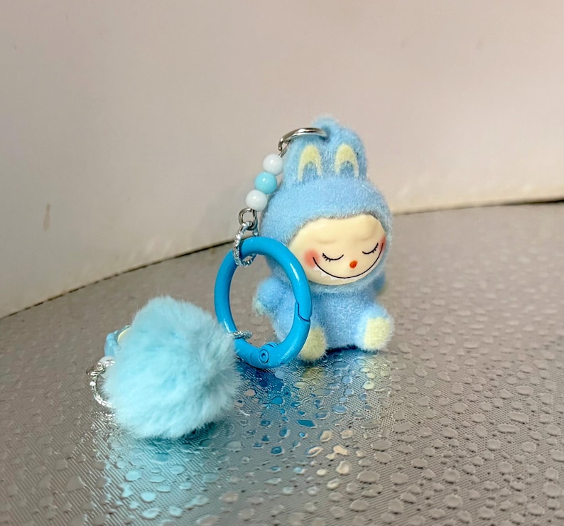 Peut inclure: Un porte-cl&eacute;s en peluche bleu clair repr&eacute;sentant un personnage de dessin anim&eacute; en costume de lapin. Le personnage a un visage souriant avec les yeux ferm&eacute;s et des accents jaunes. Un pompon bleu duveteux et un anneau bleu sont attach&eacute;s au porte-cl&eacute;s.