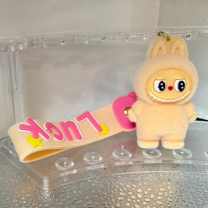 Peut inclure: Un porte-cl&eacute;s p&ecirc;che avec un personnage de dessin anim&eacute; en costume de lapin, un visage souriant et de grands yeux. Le porte-cl&eacute;s comprend une lani&egrave;re avec le mot "Luck" en lettres roses et des &eacute;toiles jaunes.
