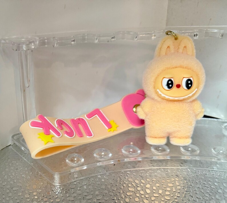 Peut inclure: Un porte-cl&eacute;s p&ecirc;che avec un personnage de dessin anim&eacute; en costume de lapin, un visage souriant et de grands yeux. Le porte-cl&eacute;s comprend une lani&egrave;re avec le mot "Luck" en lettres roses et des &eacute;toiles jaunes.