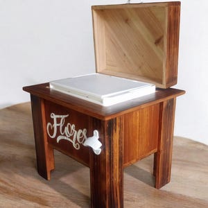 May include: A wooden box with a hinged lid, resting on a small table. The table and box are stained a warm brown. The word "Flores" is written on the side of the table in a cursive font. A white bottle opener is attached to the side.