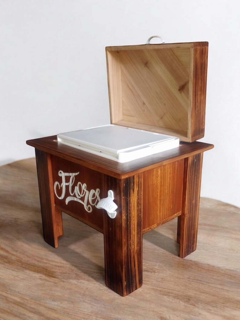 May include: A wooden box with a hinged lid, resting on a small table. The table and box are stained a warm brown. The word "Flores" is written on the side of the table in a cursive font. A white bottle opener is attached to the side.