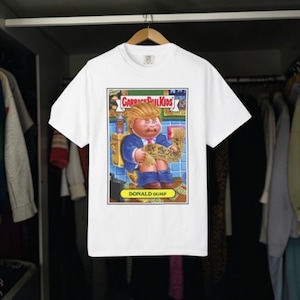 May include: White t-shirt featuring a Garbage Pail Kids parody illustration of a person on a toilet, holding a document that reads "We the People." The text "DONALD DUMP" is at the bottom of the image.