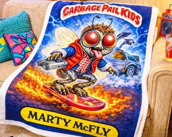 May include: A plush blanket featuring a Garbage Pail Kids character skateboarding, with the text "Marty McFly." The blanket is on a couch with retro decor, including a vintage TV, Back to the Future poster, and Teenage Mutant Ninja Turtles poster. The words "Pure Nostalgia" are at the bottom.