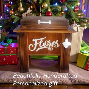 May include: A handcrafted wooden cooler with a matching lid, featuring the name "Flores" in white script. Includes a bottle opener on the side. The text "Beautifully Handcrafted Personalized gift" is at the bottom. The cooler is brown and sits in front of a Christmas tree.