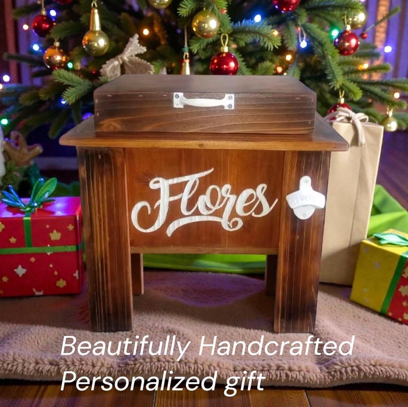 May include: A handcrafted wooden cooler with a matching lid, featuring the name "Flores" in white script. Includes a bottle opener on the side. The text "Beautifully Handcrafted Personalized gift" is at the bottom. The cooler is brown and sits in front of a Christmas tree.