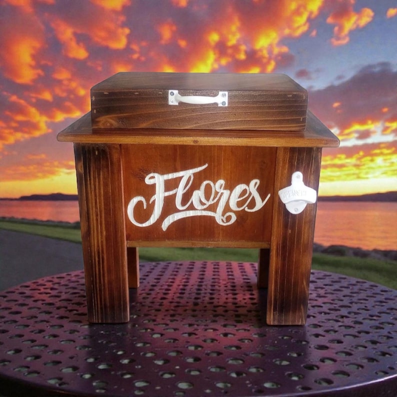 May include: A wooden cooler with a dark stain, featuring the word "Flores" in white script. It has a small, matching lid with a handle and a bottle opener on the side. The cooler is set on a metal table with a sunset backdrop.