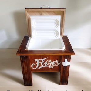 May include: A wooden cooler with a hinged lid and a white interior. The cooler is stained a dark brown and features the word "Flores" in white script. A bottle opener is attached to the side. The text "Beautifully Handmade" is at the bottom.