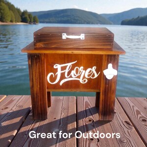 May include: A wooden cooler with a dark stain, featuring the name "Flores" in white script. It has a bottle opener on the side and a lid with a handle. The text "Great for Outdoors" is at the bottom.