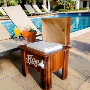 May include: A wooden cooler with a hinged lid, sitting poolside. The cooler has a white interior and the word "Flores" in cursive script on the side. A bottle opener is attached. Two cocktails and a potted succulent are nearby.