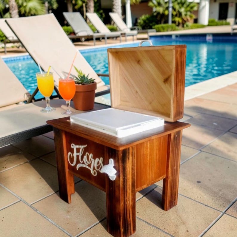 May include: A wooden cooler with a hinged lid, sitting poolside. The cooler has a white interior and the word "Flores" in cursive script on the side. A bottle opener is attached. Two cocktails and a potted succulent are nearby.