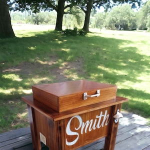 May include: A wooden cooler with a matching lid and a metal handle. The cooler is stained a warm brown and has the name "Smith" in white script on the front. A bottle opener is attached to the side.