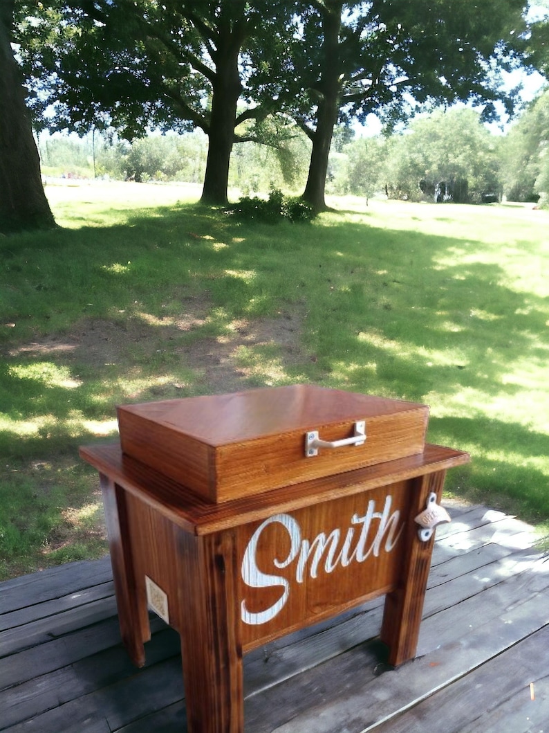 May include: A wooden cooler with a matching lid and a metal handle. The cooler is stained a warm brown and has the name "Smith" in white script on the front. A bottle opener is attached to the side.