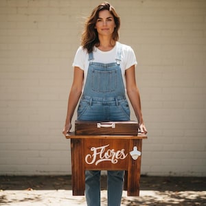 May include: A wooden cooler with the word "Flores" in white script. The cooler has a bottle opener on the side and a wooden box on top. The person in the image is wearing denim overalls and a white t-shirt.