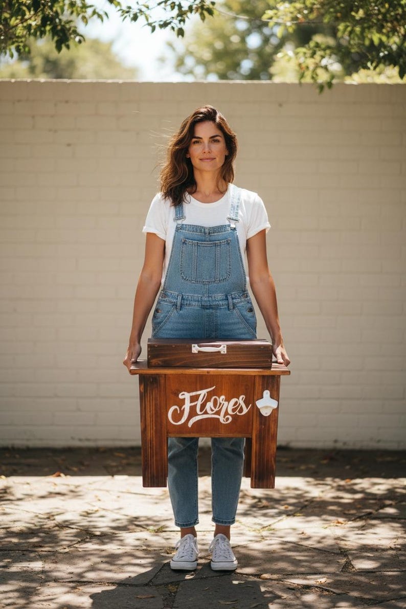 May include: A wooden cooler with the word "Flores" in white script. The cooler has a bottle opener on the side and a wooden box on top. The person in the image is wearing denim overalls and a white t-shirt.