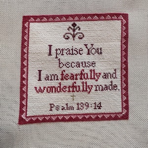 May include: A square cross-stitch embroidery on light beige fabric featuring a dark red border with a repeating triangular pattern. The text reads: I praise You because I am fearfully and wonderfully made. Psalm 139:14, with a small cross and floral motif.