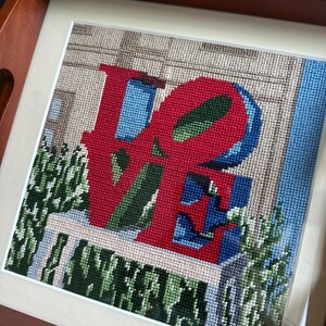 以下が含まれることがあります： 赤い文字で「LOVE」と書かれた、青と緑のアクセントが施された額入りのニードルポイントアート。 木製のトレイに、建物と緑を背景に展示されています。