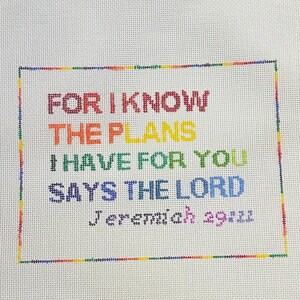 May include: A cross-stitched artwork on white fabric. The text reads "FOR I KNOW THE PLANS I HAVE FOR YOU SAYS THE LORD Jeremiah 29:11" in a rainbow of colors. The text is framed by a multi-colored border.
