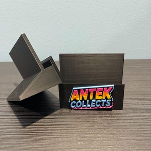 May include: A black 3D-printed display stand with a slanted back and a rectangular base. The base features the text "ANTEK COLLECTS" in a colourful, comic-book style font.