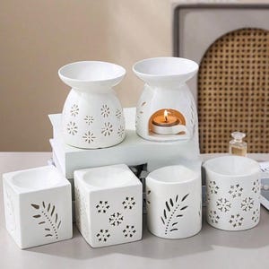 May include: A collection of white ceramic essential oil diffusers and candle holders. The diffusers have cut-out floral designs, and one is lit with a candle. The candle holders are cube and cylinder shaped with leaf and floral cut-out patterns.