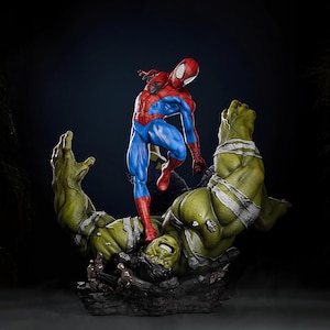 Könnte beinhalten: Detaillierte Statue von Spider-Man in seinem klassischen rot-blauen Anzug, der über einem besiegten Hulk steht. Der Hulk ist grün mit weißen Bandagen. Die Basis ist ein dunkles, felsiges Gelände.