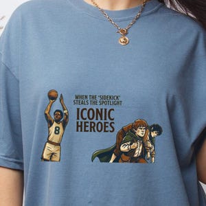 May include: A dusty blue t-shirt featuring a graphic print of a basketball player and two fantasy characters. The text reads: WHEN THE 'SIDEKICK' STEALS THE SPOTLIGHT ICONIC HEROES. The shirt has a crew neckline and short sleeves.